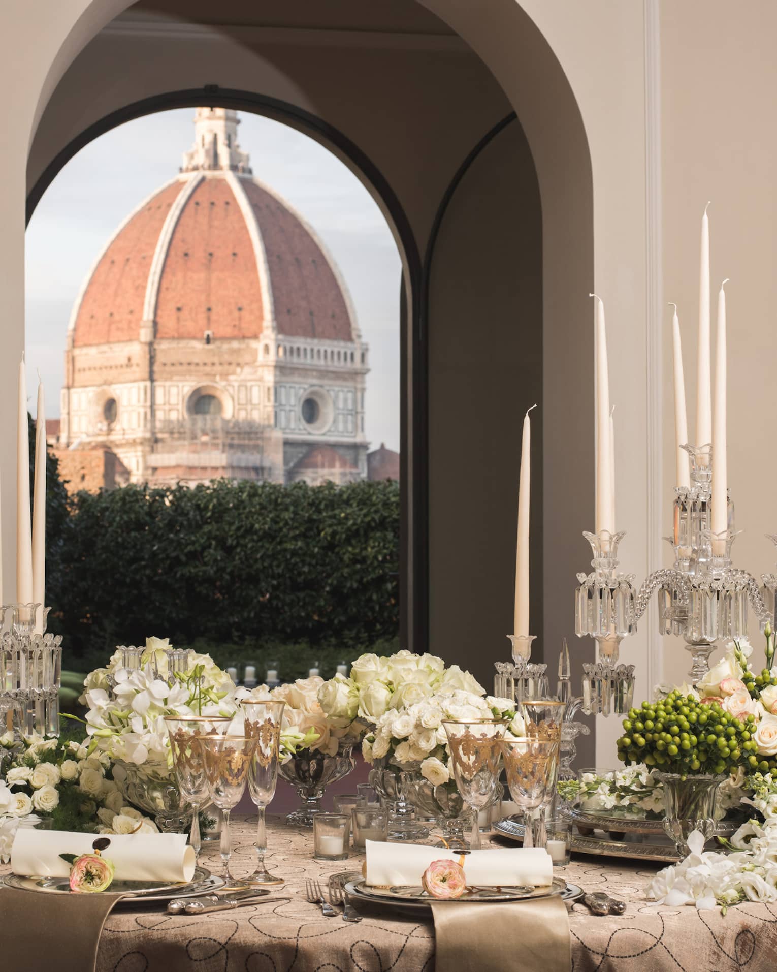 Elegant dining table set with candles, flowers, and glassware, with a large dome visible through arched windows in the background.